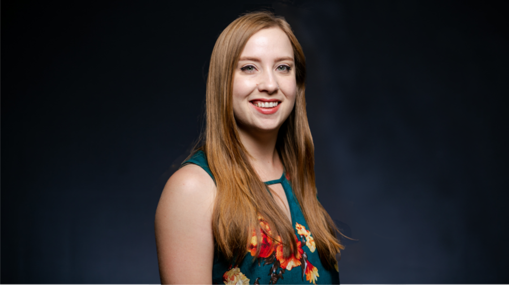 Professional headshot of a smiling woman with long hair wearing a floral top against a dark background